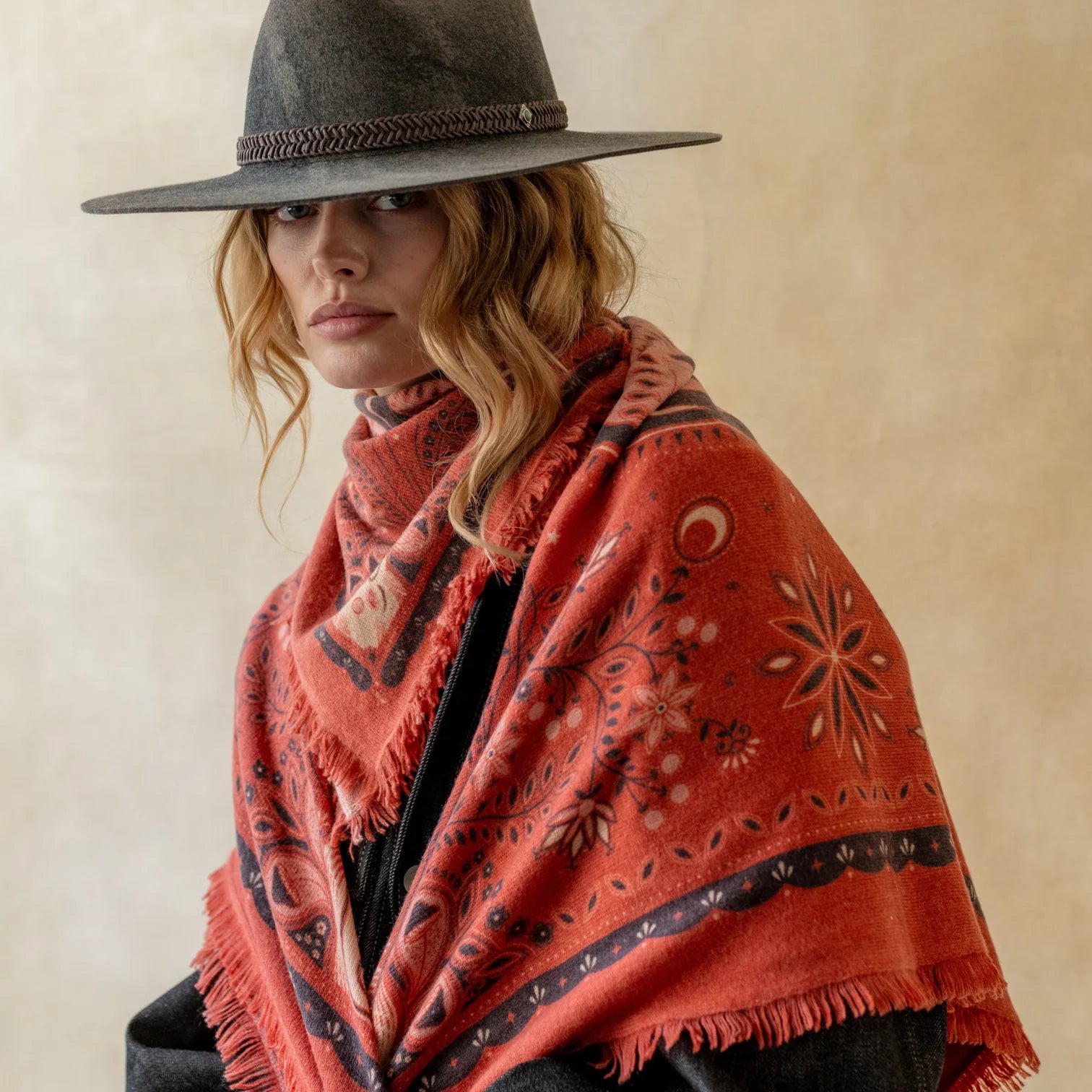 A woman wearing a wide-brimmed dark hat and a rust-orange patterned shawl with fringe, photographed against a warm beige studio background.
