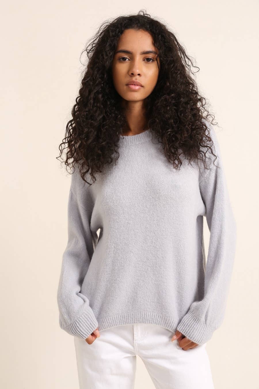 Woman wearing a light gray sweater and white pants against a beige background