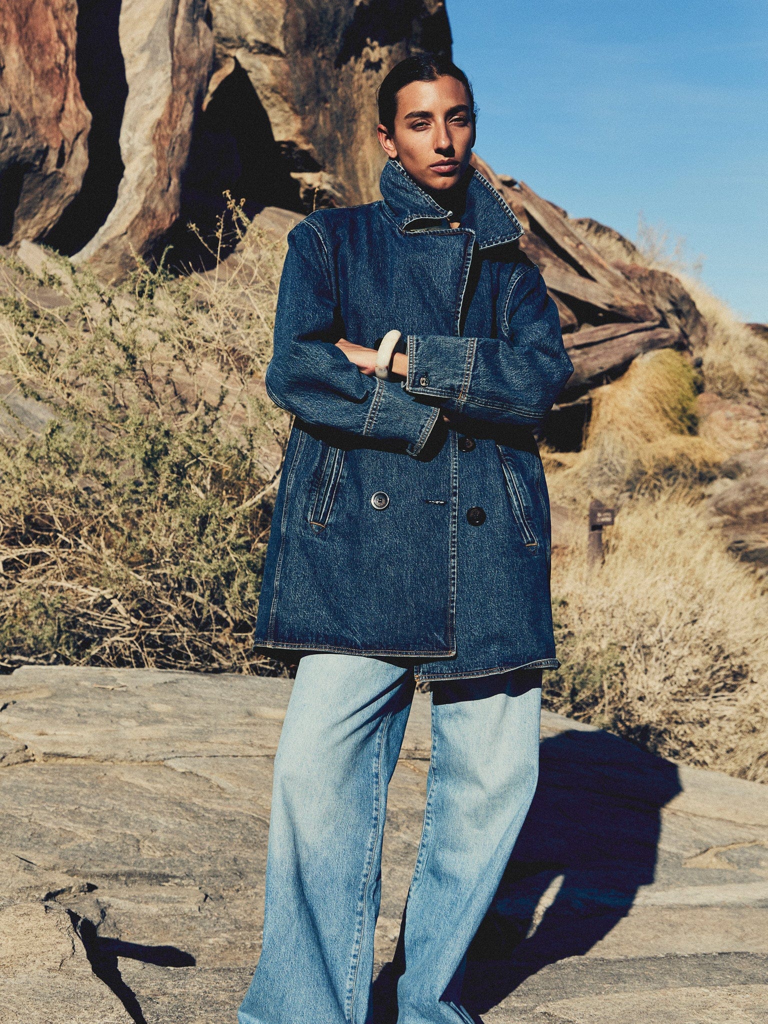 Person wearing a denim jacket and jeans standing in a rocky outdoor setting.